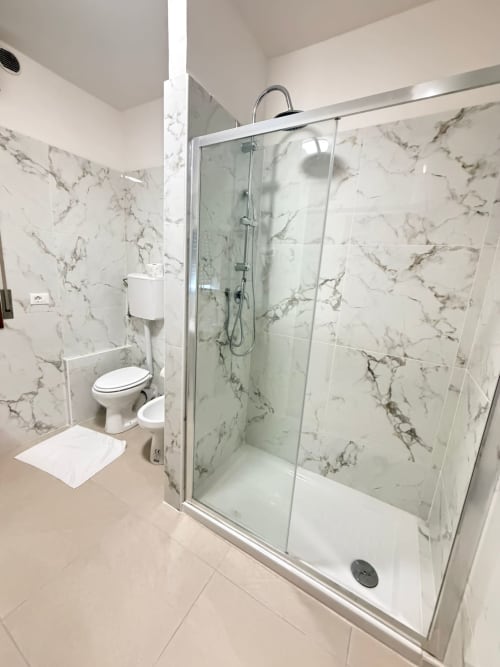 Bagno moderno con rivestimenti effetto marmo, box doccia in vetro, wc e pavimento bianco presso TMS Hotel Mestre