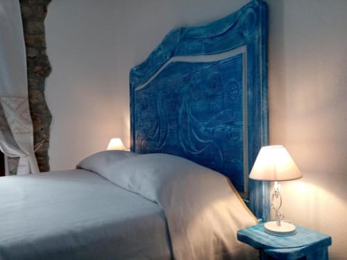 Camera elegante a Regnos Altos Bosa con testata blu intagliata, biancheria bianca, lampada comodino e travi rustica