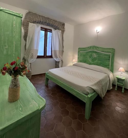 Camera luminosa con letto in legno verde, biancheria beige e fiori freschi presso Regnos Altos Boutique Rooms a Bosa