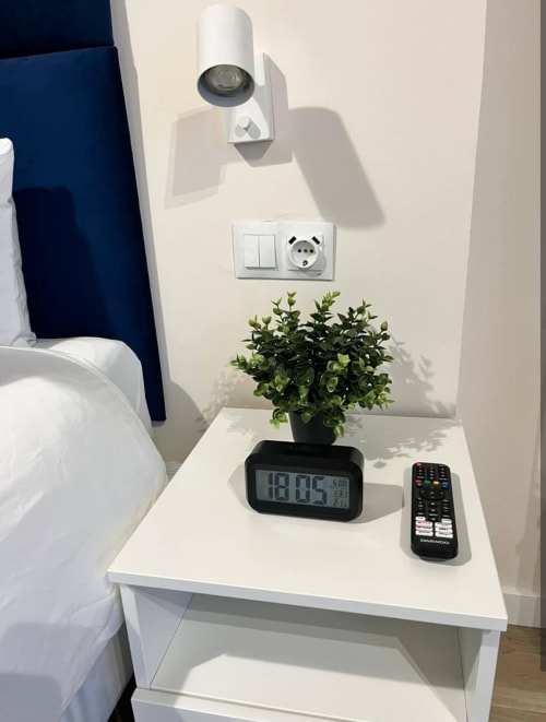 Mesita de noche en habitación de hotel Valencia con reloj digital, control remoto y planta en maceta junto a la cama