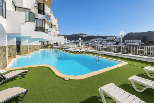 Terraço da piscina Apartamentos Gelimar com espreguiçadeiras, vista do mar e edifícios das colinas de Puerto Rico sob céu azul