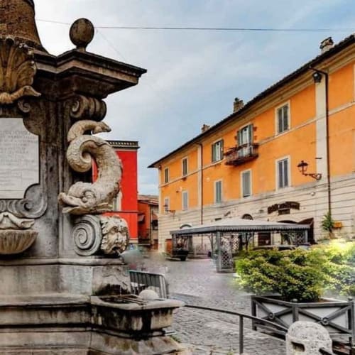 Piazza storica italiana presso Travellers Homes a Campagnano di Roma con fontana in pietra ornamentale e facciata dell'edificio arancione