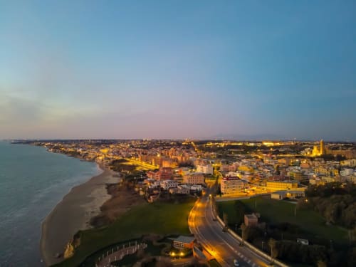 Cittadina costiera italiana al tramonto con edifici illuminati, spiaggia e strada lungomare a Campagnano di Roma
