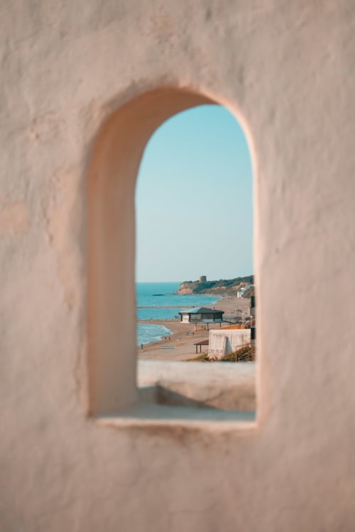 Finestra ad arco che incornicia la vista della spiaggia mediterranea con acque turchesi e scogliere costiere vicino a Roma