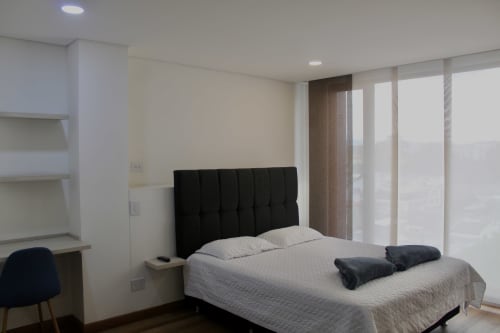 Dormitorio moderno en apartaestudio Modelia Living, Bogotá, con cabecera negra, vista a la ciudad, escritorio y TV de pantalla plana