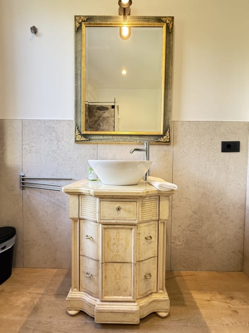 Elegante lavabo con vasca bianca su vanità in legno crema, specchio con cornice dorata, pareti piastrellate e illuminazione calda