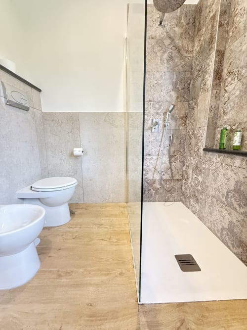 Bagno moderno con doccia walk-in, toilette e bidet presso B&B Luna Verde in Italia. Rivestimenti in travertino e pavimento in legno.