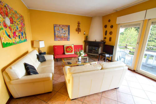 Luminoso soggiorno giallo nella villa di Lake Orta con divani color crema, divano rosso accento, camino e balcone con vista sul lago