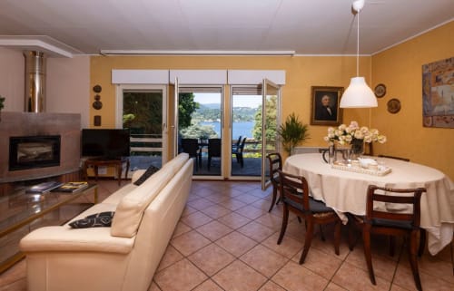Ampia zona living con divano color panna, tavolo da pranzo, camino e vista sul lago d'Orta con accesso alla terrazza esterna