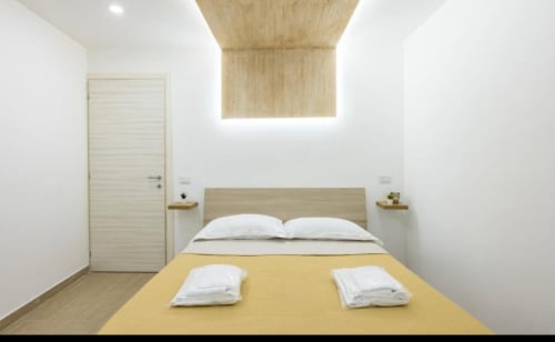 Camera moderna a Casa Fernanda a Napoli con letto in legno, cuscini bianchi, coperta gialla e plafoniera contemporanea