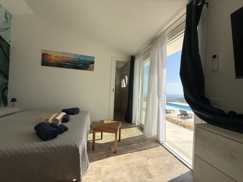 Bright bedroom with double bed, direct pool access via glass doors, ocean view, TV, and modern amenities at Sardinia Holidays