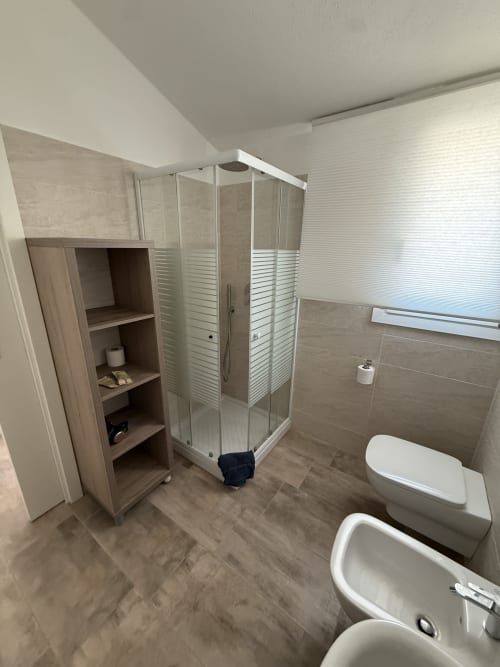 Modern bathroom at Sardinia Holidays with glass shower enclosure, toilet, bidet, and wooden storage cabinet