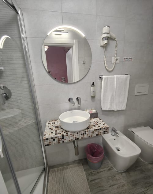 Bagno moderno dell'Hotel Principe Amedeo con specchio rotondo, lavabo bianco, doccia e piastrelle mosaico