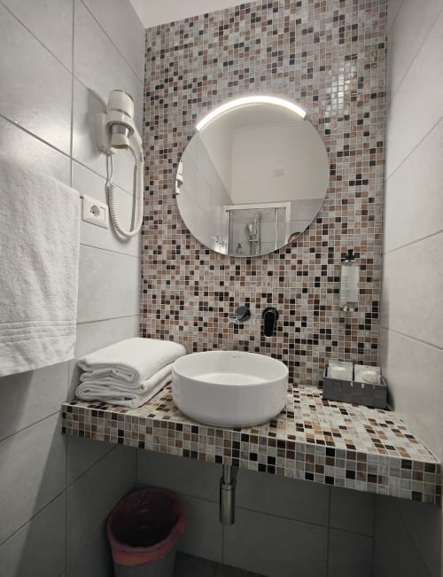 Bagno moderno dell'Hotel Principe Amedeo con specchio rotondo, lavabo bianco, piastrelle a mosaico e servizi a Roma