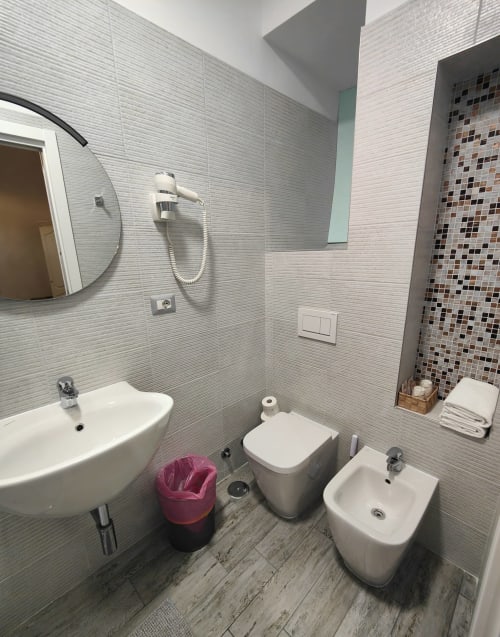 Bagno moderno dell'Hotel Principe Amedeo con lavabo bianco, bidet, toilette, piastrelle grigie e accessori doccia