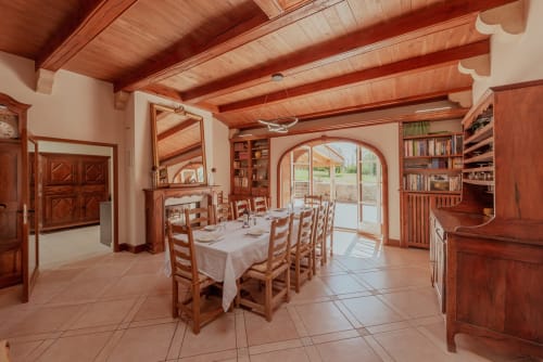 Élégante salle à manger de la villa Vila Cabanes 46 dans le Lot, avec poutres apparentes, chaises rustiques, cheminée et portes-fenêtres en arche donnant sur le jardin