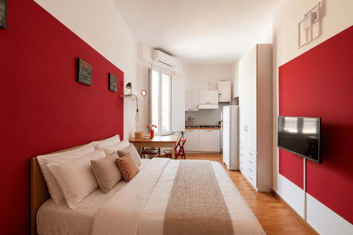 Modern studio apartment in Milano's Navigli district with red accent walls, wooden flooring, bed, kitchenette, and