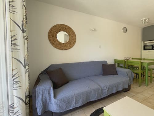 Séjour de l'appartement Vallon des Sources avec canapé bleu, miroir rond tressé et coin repas à Digne-les-Bains