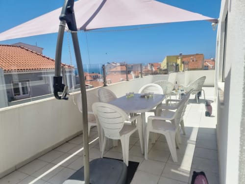 Terraço na cobertura da Casa da Barroca em Ericeira com mesa de refeições, cadeiras brancas e vistas da cidade sob guarda-sol