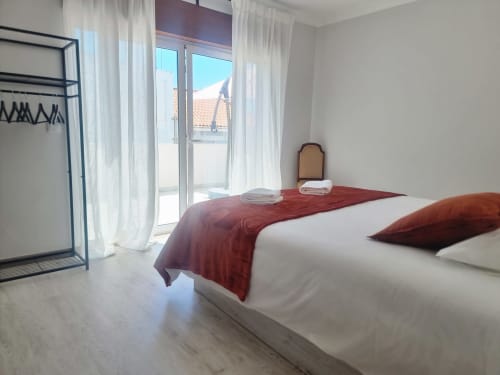 Quarto luminoso na Casa da Barroca em Ericeira com cama de casal, manta vermelha, vista da cidade pela porta de vidro e design minimalista moderno