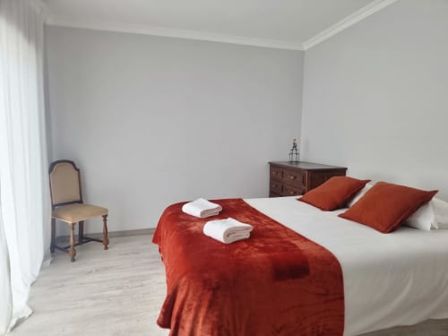 Quarto luminoso da Casa da Barroca, Ericeira com roupa de cama vermelha, almofadas brancas, cómoda em madeira e vista da cidade