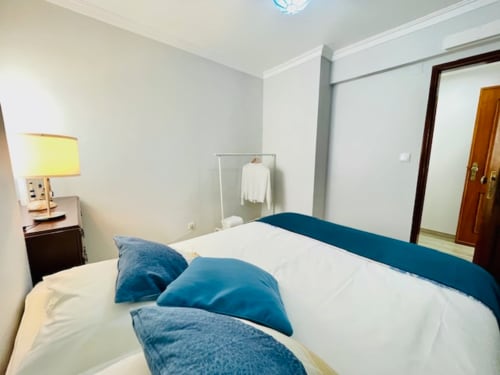 Quarto luminoso na Casa da Barroca em Ericeira com almofadas azuis, roupa de cama branca, luminária e porta de madeira escura