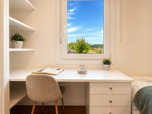 Escritorio luminoso con vista al mar hacia Baiona, España. Espacio beige con estantes, planta, taza de café y libro abierto