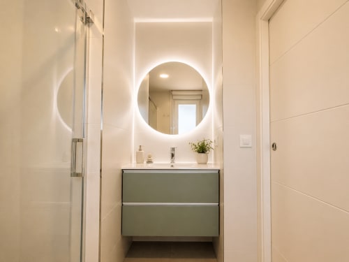 Baño moderno en apartamento de Baiona con espejo redondo iluminado, tocador verde, paredes de azulejos blancos y cabina de ducha