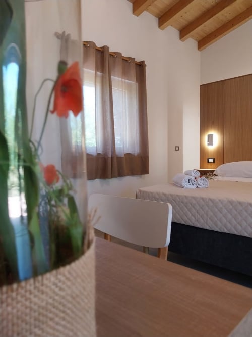 Modern hotel room at Agriturismo Lumia Resort with queen bed, wooden beams, brown curtains, and red flowers on nightstand