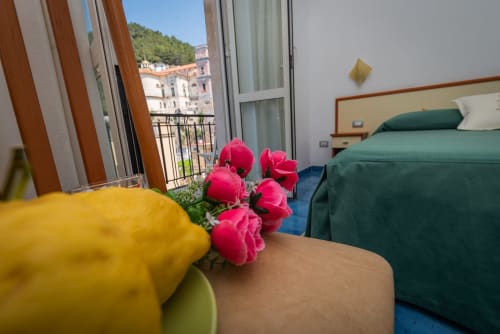 Luminosa camera d'hotel con fiori rosa, letto verde e balcone che affaccia sull'architettura storica di Maiori