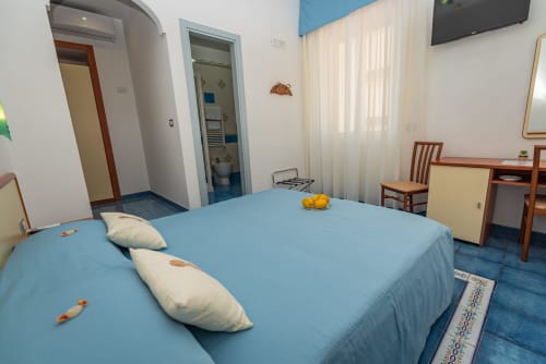 Ampia camera da letto a Casa Mannini B&B a Maiori con biancheria blu, cuscini bianchi, mobili in legno e bagno privato
