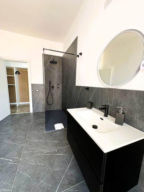 Bagno moderno dell'Hotel Newhouse a Bari con piastrelle in marmo scuro, lavabo bianco, specchio rotondo e doccia a filo pavimento