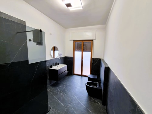 Bagno moderno nella suite Newhouse Cavour Deluxe a Bari. Piastrelle di marmo nero, pareti bianche, lavabo, wc e porte in legno.