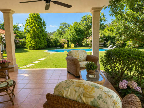 Patio cubierto con sillas de mimbre con vistas al frondoso jardín y piscina azul de la villa Holibai en Tomiño. Galicia
