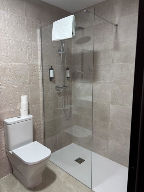 Baño moderno del Hotel Mulhacén Trevélez con inodoro blanco, mampara de ducha de cristal y azulejos texturados en tono beige