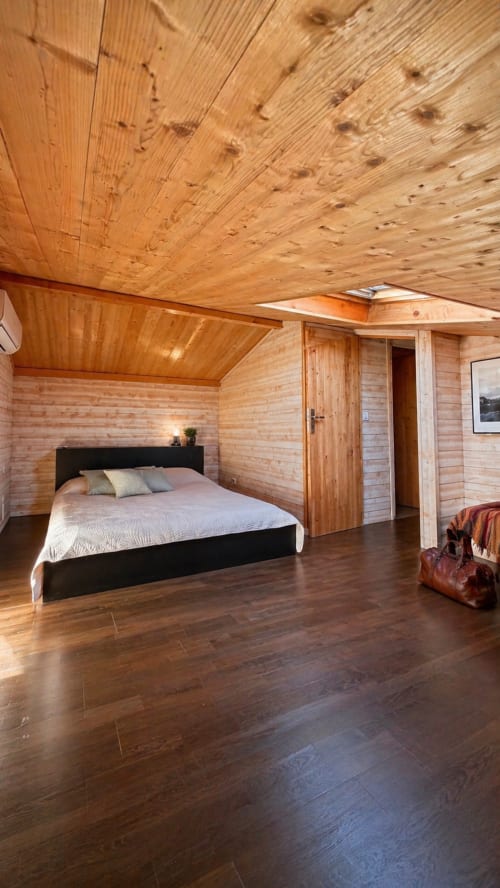 Chambre spacieuse de la villa en bois au Maquis Plage avec lit noir, plafond en bois, sol foncé et équipements modernes