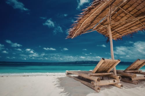 Wooden beach lounge chairs under thatch umbrella facing turquoise ocean and white sand beach