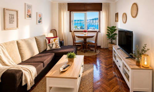Luminoso salón en Baiona con vistas al mar, sofá marrón, muebles de madera, televisor y puerto costero visible