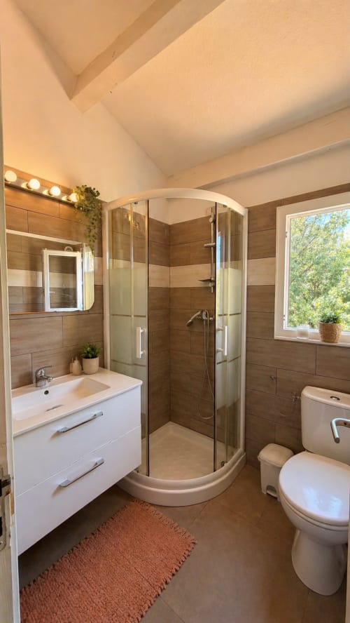 Salle de bains moderne avec murs en bois-carrelage, vasque blanche, douche d'angle, toilettes et fenêtre à la villa Le Maquis Plage