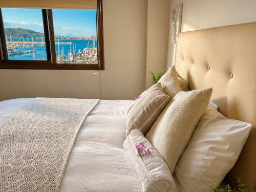 Dormitorio luminoso con cabecero crema, ventanas con vistas al mar sobre la marina de Baiona y veleros, ropa de cama y almohadas beige