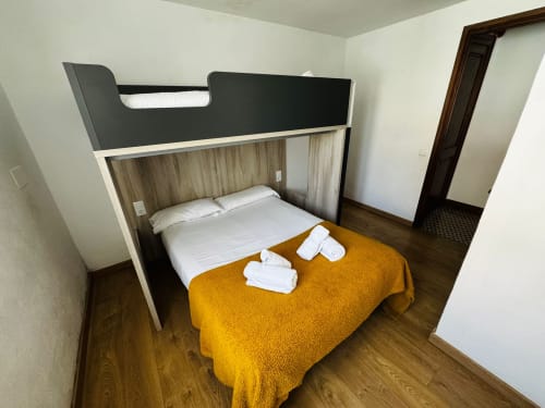 Dormitori modern a la vila El Niu de l'Aguila, Camprodon, amb llit encastat, manta groga, tovalloles blanques i sòl de fusta