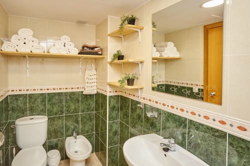 Baño moderno en Experiencias Encant de Calafat con azulejos verdes, sanitarios blancos y estantes de madera con toallas