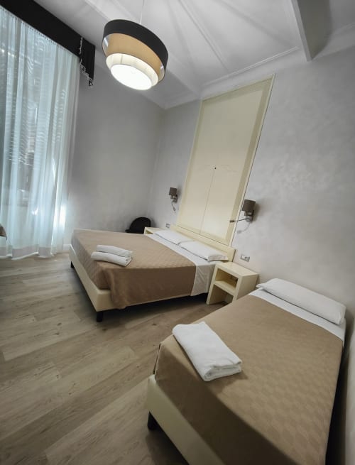 Moderna camera d'hotel Principe Amedeo con due letti, pavimenti in legno, illuminazione a soffitto, grande specchio e finestra con tende