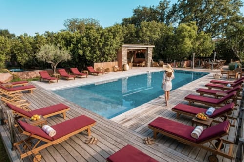 Piscine de la station balnéaire Le Maquis Plage avec transats bordeaux, terrasse en bois et pavillon en pierre entouré d'arbres méditerranéens