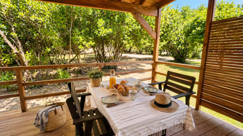 Shaded wooden terrace at Le Maquis Plage with table setting, bread, and hat overlooking green gardens