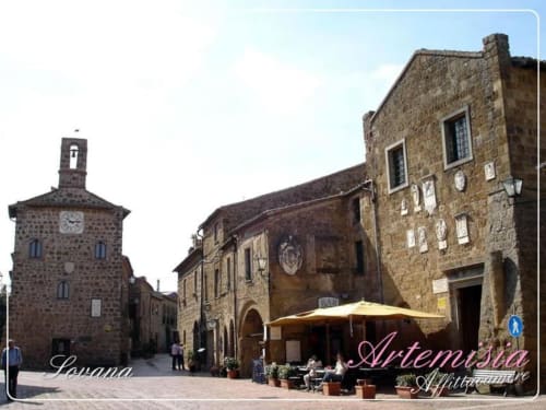 Piazza medievale di Capalbio con edifici in pietra, torre dell'orologio e ombrelloni del caffè del B&B Casale Artemisia