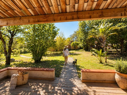 Pergolato in legno sulla terrazza dell'hotel Le Maquis Plage a Poggio-Mezzana con vista sui rigogliosi giardini, una donna e il suo cane lungo il sentiero
