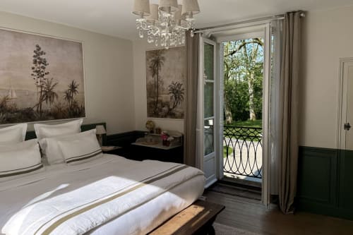 Elegant bedroom at La Maison 1680 with garden views, crystal chandelier, botanical artwork, and French doors opening to
