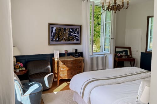 Elegant bedroom at La Maison 1680 with bed, leather chair, wooden dresser, and garden views through French doors