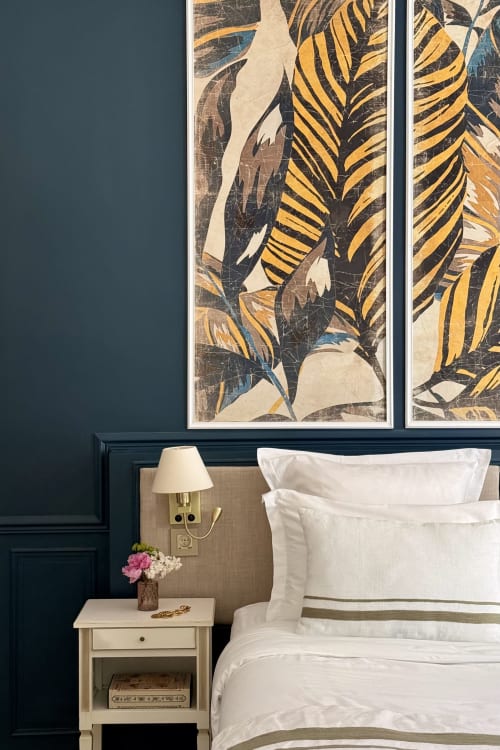 Elegant bedroom at La Maison 1680 with botanical framed art, white bedding, navy walls, and cream nightstand with lamp.